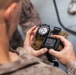 A military personnel in desert camouflage uniform is holding a combined navigation device consisting of a magnetic compass mounted on top of a small GPS/digital device with a tan MOLLE-style strap. The device displays a menu screen with options including 'DELETE MAP' and 'OPTIONS', along with coordinate data. The image is taken from an over-the-shoulder perspective near what appears to be a military vehicle.