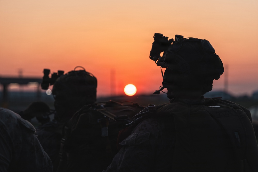 Two military personnel are silhouetted against a vivid orange and red sunset sky. Both figures are equipped with tactical gear including helmets mounted with night vision devices (NVDs) and appear to be carrying backpacks or rucksacks. The image is a dramatic low-light photograph emphasizing the contrast between the dark silhouettes and the glowing horizon.