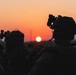 Two military personnel are silhouetted against a vivid orange and red sunset sky. Both figures are equipped with tactical gear including helmets mounted with night vision devices (NVDs) and appear to be carrying backpacks or rucksacks. The image is a dramatic low-light photograph emphasizing the contrast between the dark silhouettes and the glowing horizon.