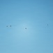 The image shows five parachutists descending through a clear blue sky. The parachutes appear small and distant, suggesting the subjects are at considerable altitude. The minimalist composition features only sky and the parachutists, with no ground or horizon visible.