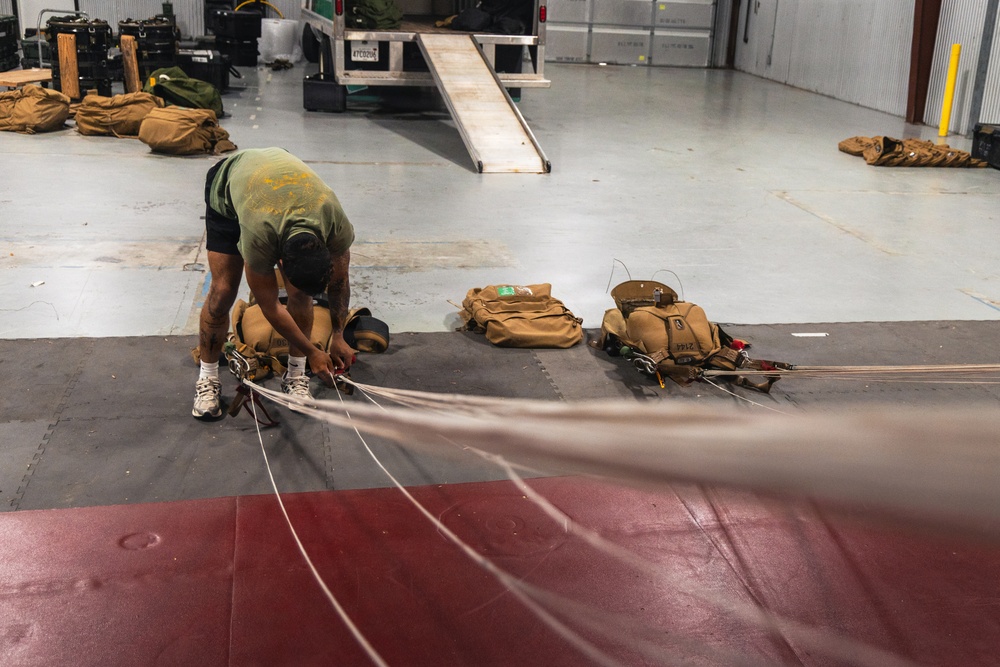 A person in a green t-shirt and black shorts is bent over working with parachute equipment on a large mat inside what appears to be a military or aviation hangar. Multiple tan/olive-colored parachute packs and rigging lines are laid out on the floor in an organized manner. A truck with a loading ramp is visible in the background along with additional equipment bags.