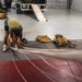 A person in a green t-shirt and black shorts is bent over working with parachute equipment on a large mat inside what appears to be a military or aviation hangar. Multiple tan/olive-colored parachute packs and rigging lines are laid out on the floor in an organized manner. A truck with a loading ramp is visible in the background along with additional equipment bags.