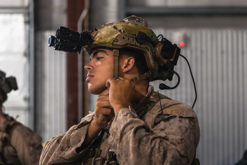 A military service member is adjusting the chin strap of a tactical helmet in what appears to be an indoor facility. The individual is wearing full combat gear including a multicam helmet mounted with night vision goggles and communication headset. The uniform pattern visible is consistent with USMC MARPAT desert camouflage.