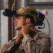 A military service member is adjusting the chin strap of a tactical helmet in what appears to be an indoor facility. The individual is wearing full combat gear including a multicam helmet mounted with night vision goggles and communication headset. The uniform pattern visible is consistent with USMC MARPAT desert camouflage.