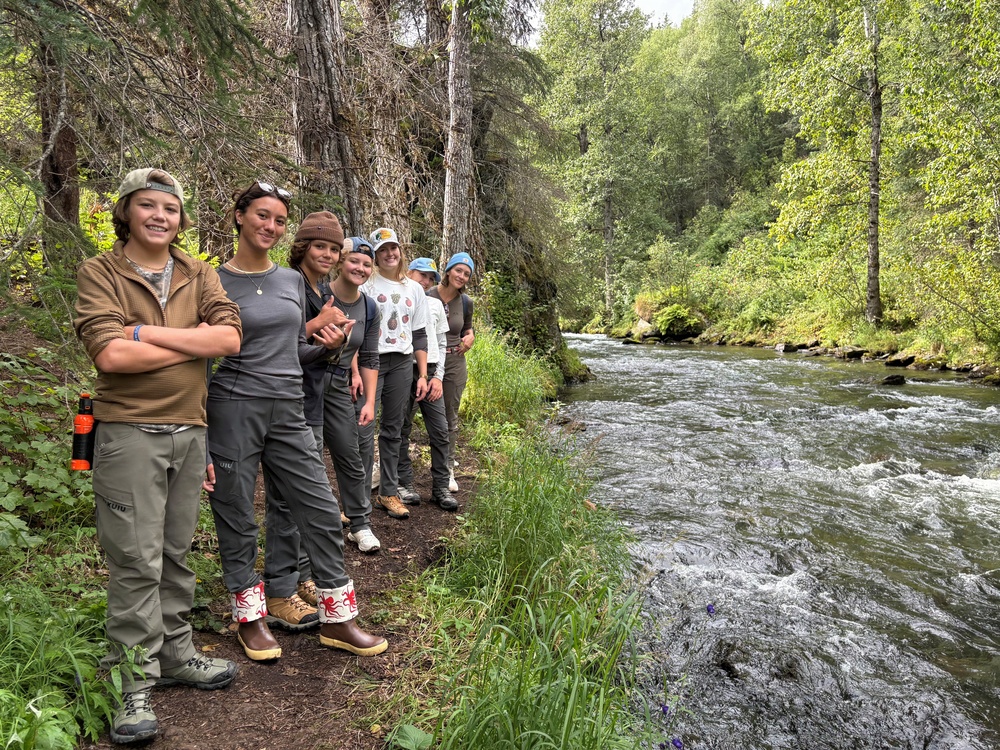 A group of approximately seven young people pose together along a forested riverbank trail, smiling at the camera. They are dressed in outdoor/hiking attire including cargo pants, hiking boots, and hats, suggesting a group hiking or outdoor education excursion. A clear, fast-moving stream runs alongside them, bordered by dense green forest.