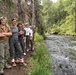 A group of approximately seven young people pose together along a forested riverbank trail, smiling at the camera. They are dressed in outdoor/hiking attire including cargo pants, hiking boots, and hats, suggesting a group hiking or outdoor education excursion. A clear, fast-moving stream runs alongside them, bordered by dense green forest.