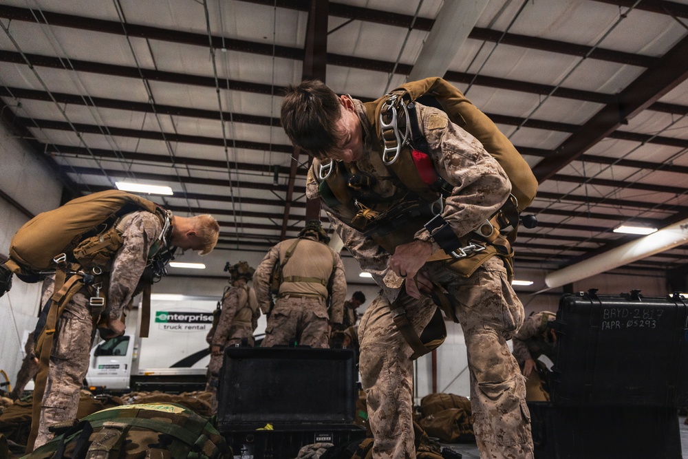 Military personnel in desert camouflage uniforms are inside a large warehouse or hangar, preparing parachute and jump equipment. Multiple service members are bent over inspecting or adjusting their harnesses and gear. An Enterprise truck rental vehicle is visible in the background through an opening.