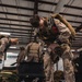 Military personnel in desert camouflage uniforms are inside a large warehouse or hangar, preparing parachute and jump equipment. Multiple service members are bent over inspecting or adjusting their harnesses and gear. An Enterprise truck rental vehicle is visible in the background through an opening.