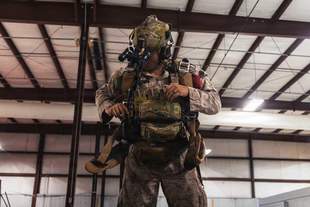 A heavily equipped military service member stands inside what appears to be a large hangar or warehouse facility. The individual is wearing MARPAT (Marine pattern) desert camouflage uniform with extensive tactical gear including a helmet-mounted night vision device, plate carrier, and parachute or jump harness. The soldier appears to be conducting a gear check or pre-mission equipment inspection.