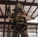 A heavily equipped military service member stands inside what appears to be a large hangar or warehouse facility. The individual is wearing MARPAT (Marine pattern) desert camouflage uniform with extensive tactical gear including a helmet-mounted night vision device, plate carrier, and parachute or jump harness. The soldier appears to be conducting a gear check or pre-mission equipment inspection.