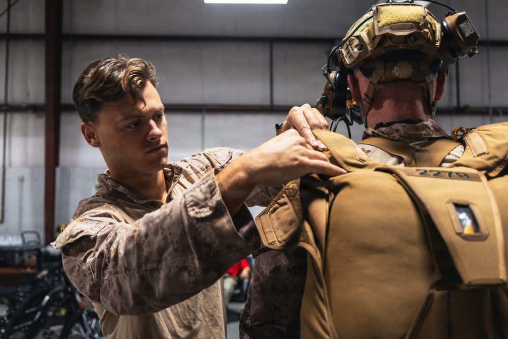 Two military personnel in desert camouflage uniforms are shown inside what appears to be a hangar or equipment bay. One service member is assisting the other by adjusting or securing tactical gear on the back of a fully equipped operator. The equipped individual is wearing a tactical helmet with communications headset and night vision mount, along with a tan plate carrier or parachute rig marked with the number '2279'.