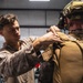 Two military personnel in desert camouflage uniforms are shown inside what appears to be a hangar or equipment bay. One service member is assisting the other by adjusting or securing tactical gear on the back of a fully equipped operator. The equipped individual is wearing a tactical helmet with communications headset and night vision mount, along with a tan plate carrier or parachute rig marked with the number '2279'.