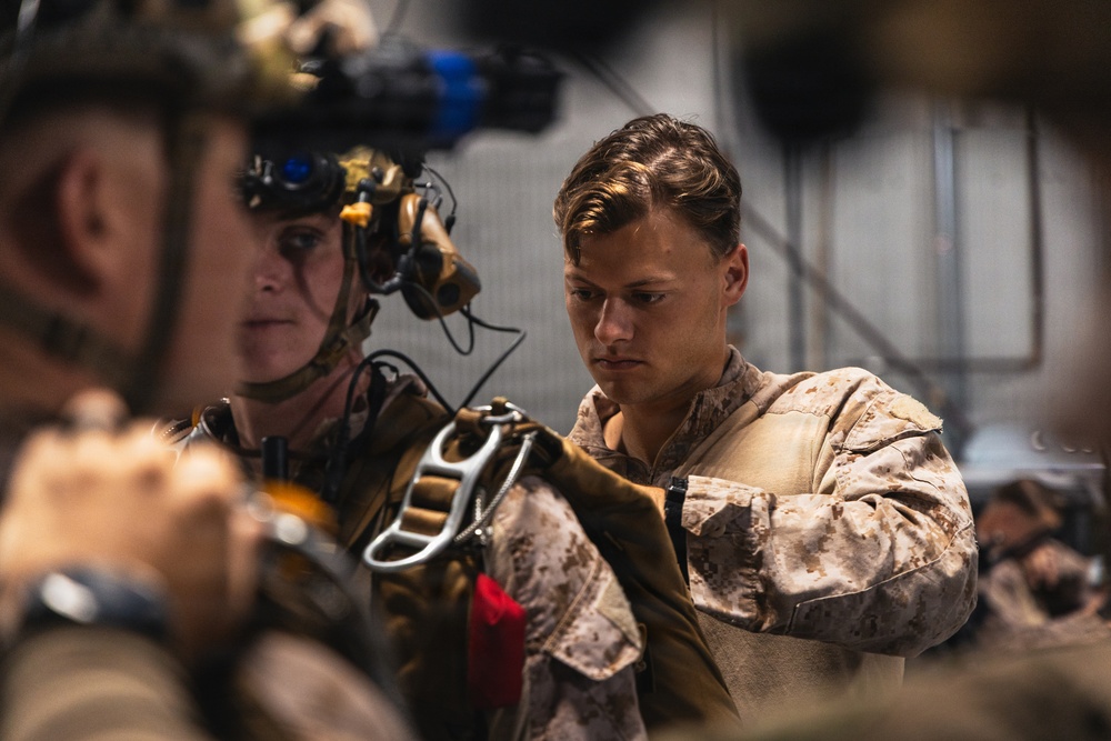 Two military personnel in desert camouflage (MARPAT) uniforms are engaged in pre-mission equipment checks. One individual is wearing a tactical helmet equipped with night vision goggles and communications headset, while the other is inspecting or adjusting carabiner and harness equipment. The setting appears to be an indoor staging area or hangar.