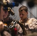 Two military personnel in desert camouflage (MARPAT) uniforms are engaged in pre-mission equipment checks. One individual is wearing a tactical helmet equipped with night vision goggles and communications headset, while the other is inspecting or adjusting carabiner and harness equipment. The setting appears to be an indoor staging area or hangar.