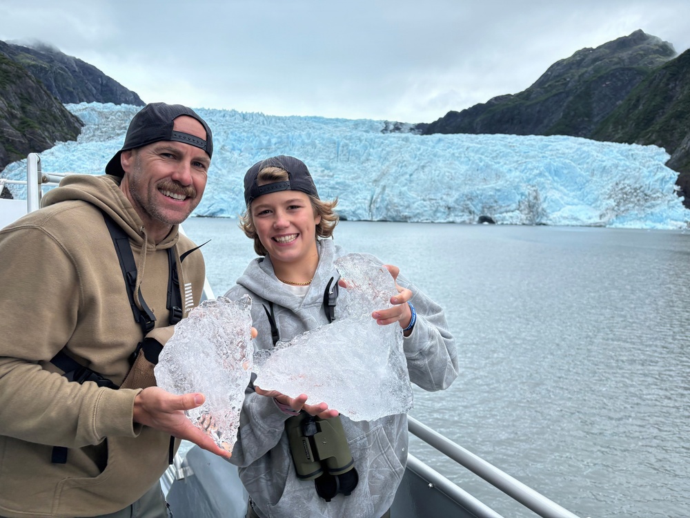 Two people, likely a father and child, stand on the deck of a tour boat holding large chunks of glacial ice. They are posing and smiling in front of a massive blue glacier flanked by rocky, vegetation-covered mountains. The setting appears to be a glacial fjord, consistent with locations such as Kenai Fjords National Park in Alaska.
