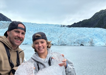 Two people, likely a father and child, stand on the deck of a tour boat holding large chunks of glacial ice. They are posing and smiling in front of a massive blue glacier flanked by rocky, vegetation-covered mountains. The setting appears to be a glacial fjord, consistent with locations such as Kenai Fjords National Park in Alaska.