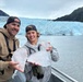 Two people, likely a father and child, stand on the deck of a tour boat holding large chunks of glacial ice. They are posing and smiling in front of a massive blue glacier flanked by rocky, vegetation-covered mountains. The setting appears to be a glacial fjord, consistent with locations such as Kenai Fjords National Park in Alaska.