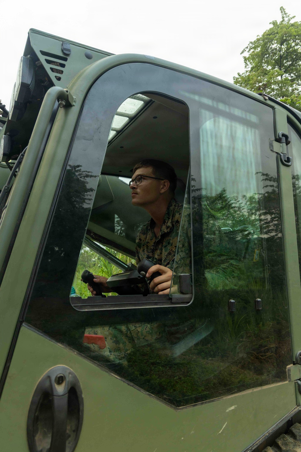 A young male service member wearing camouflage uniform is seated inside the cab of a military vehicle, viewed from a low exterior angle. He is holding what appears to be a control device or joystick and looking forward attentively. The vehicle is olive/military green and is surrounded by dense tropical vegetation.
