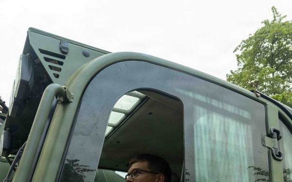 A young male service member wearing camouflage uniform is seated inside the cab of a military vehicle, viewed from a low exterior angle. He is holding what appears to be a control device or joystick and looking forward attentively. The vehicle is olive/military green and is surrounded by dense tropical vegetation.
