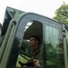 A young male service member wearing camouflage uniform is seated inside the cab of a military vehicle, viewed from a low exterior angle. He is holding what appears to be a control device or joystick and looking forward attentively. The vehicle is olive/military green and is surrounded by dense tropical vegetation.