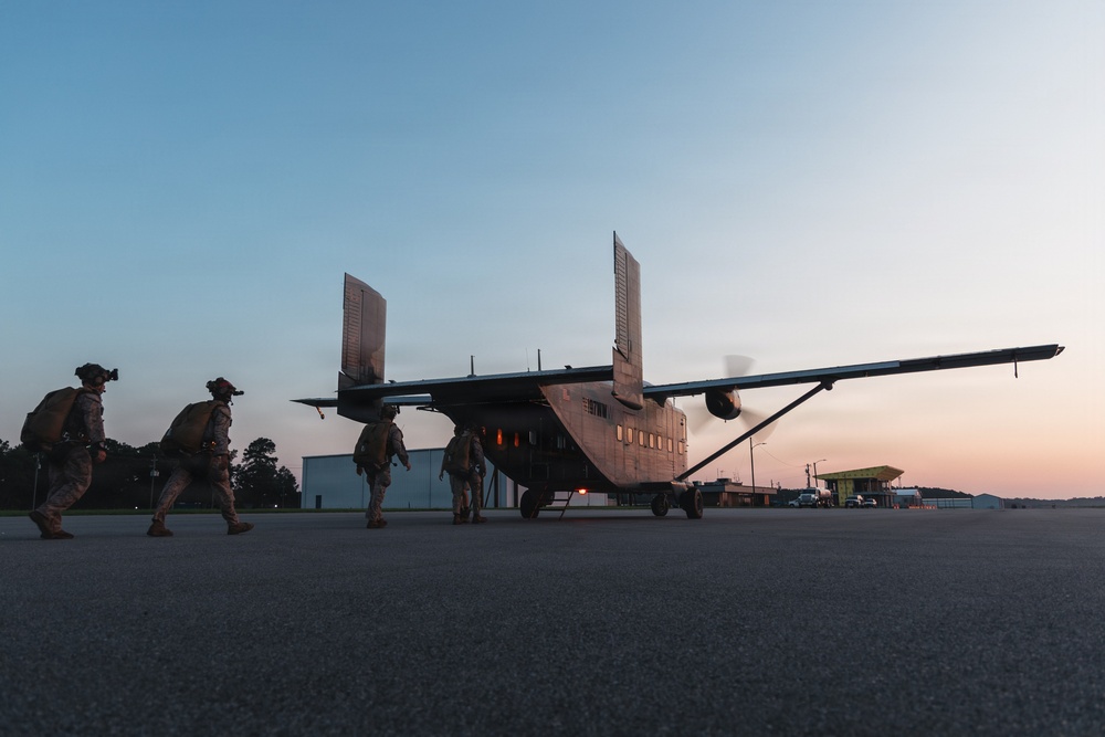 Military personnel in full combat gear and parachute equipment approach a twin-engine aircraft on a tarmac at dusk or dawn. The scene appears to depict a pre-jump or insertion operation, with soldiers boarding or preparing to board the aircraft. The low-light conditions and silhouetted figures create a dramatic operational atmosphere.