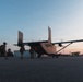 Military personnel in full combat gear and parachute equipment approach a twin-engine aircraft on a tarmac at dusk or dawn. The scene appears to depict a pre-jump or insertion operation, with soldiers boarding or preparing to board the aircraft. The low-light conditions and silhouetted figures create a dramatic operational atmosphere.