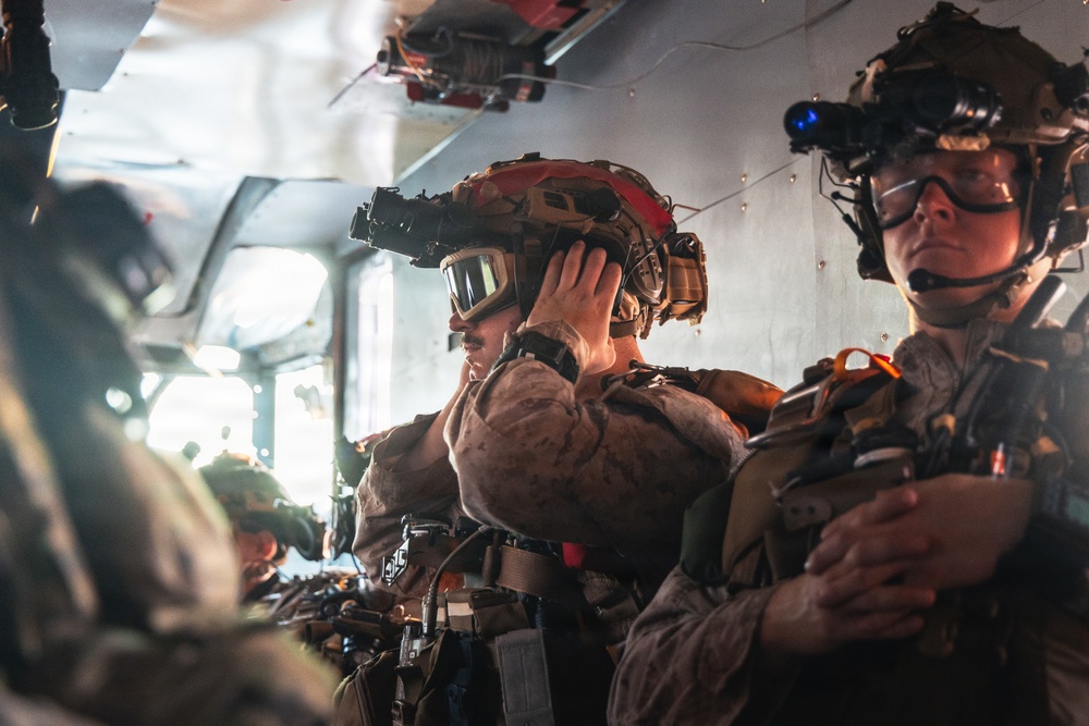 Military personnel are seated inside what appears to be a military helicopter or transport aircraft, fully equipped with tactical gear. At least two soldiers are prominently visible in the foreground, wearing helmets mounted with night vision devices (NVDs), tactical goggles, communication headsets, and body armor. The scene suggests a pre-mission or in-flight insertion scenario.