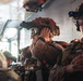 Military personnel are seated inside what appears to be a military helicopter or transport aircraft, fully equipped with tactical gear. At least two soldiers are prominently visible in the foreground, wearing helmets mounted with night vision devices (NVDs), tactical goggles, communication headsets, and body armor. The scene suggests a pre-mission or in-flight insertion scenario.
