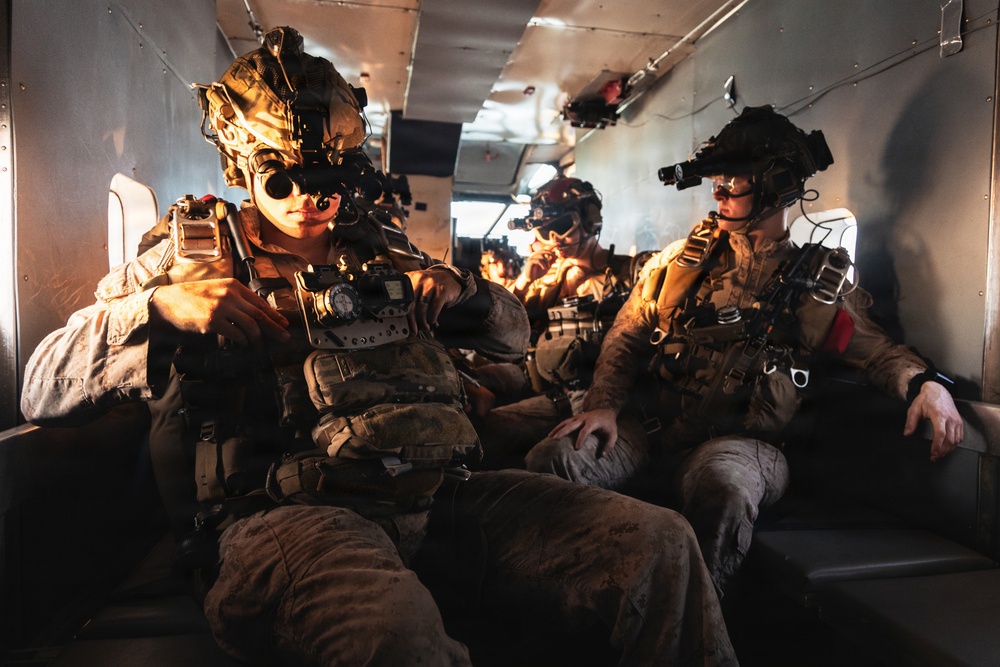 Multiple heavily equipped military personnel are seated inside what appears to be a military helicopter or transport aircraft. The soldiers are wearing full tactical gear including night vision devices mounted on their helmets, body armor, and combat uniforms. The warm, dramatic lighting suggests either a sunset/sunrise environment or artificial lighting within the aircraft cabin.