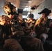 Multiple heavily equipped military personnel are seated inside what appears to be a military helicopter or transport aircraft. The soldiers are wearing full tactical gear including night vision devices mounted on their helmets, body armor, and combat uniforms. The warm, dramatic lighting suggests either a sunset/sunrise environment or artificial lighting within the aircraft cabin.
