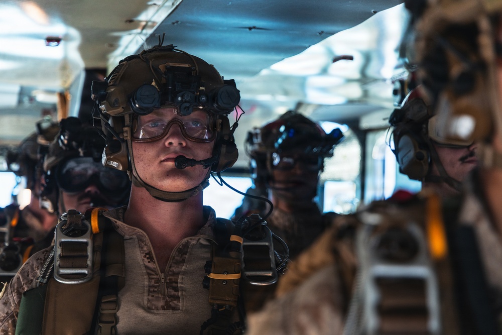A group of heavily equipped military personnel are staged inside what appears to be a military aircraft or transport vehicle. The foreground subject is wearing full tactical gear including a combat helmet with night vision device mounts, ballistic goggles, communications headset with boom microphone, and parachute harness straps. Multiple similarly equipped personnel are visible in the background, suggesting a pre-jump or pre-mission staging scenario.