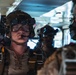 A group of heavily equipped military personnel are staged inside what appears to be a military aircraft or transport vehicle. The foreground subject is wearing full tactical gear including a combat helmet with night vision device mounts, ballistic goggles, communications headset with boom microphone, and parachute harness straps. Multiple similarly equipped personnel are visible in the background, suggesting a pre-jump or pre-mission staging scenario.