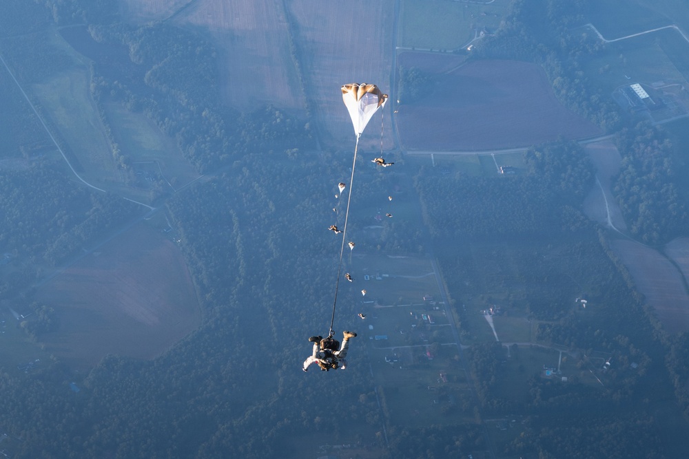 An aerial photograph captures a tandem skydiving pair in freefall with a drogue parachute deployed above them, connected by a long bridle cord. Multiple other skydivers are visible in the surrounding airspace at various distances. The image is taken from high altitude, revealing a patchwork of agricultural fields, forests, and rural landscape far below.