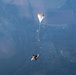 An aerial photograph captures a tandem skydiving pair in freefall with a drogue parachute deployed above them, connected by a long bridle cord. Multiple other skydivers are visible in the surrounding airspace at various distances. The image is taken from high altitude, revealing a patchwork of agricultural fields, forests, and rural landscape far below.