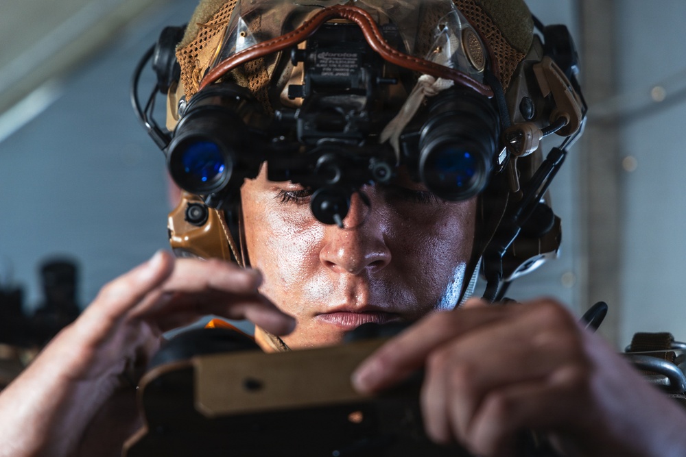 A close-up photograph of a military or special operations personnel wearing a tactical helmet mounted with dual-tube night vision goggles (NVGs) with blue-tinted lenses. The individual appears to be adjusting or operating equipment held in both hands. The image is sharply focused and professionally lit, highlighting the advanced tactical gear.