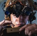 A close-up photograph of a military or special operations personnel wearing a tactical helmet mounted with dual-tube night vision goggles (NVGs) with blue-tinted lenses. The individual appears to be adjusting or operating equipment held in both hands. The image is sharply focused and professionally lit, highlighting the advanced tactical gear.