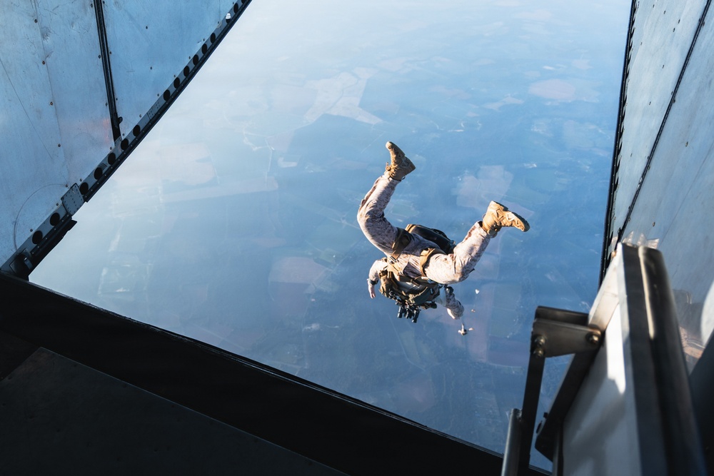 A military parachutist has just exited the rear cargo ramp of a large military aircraft, visible in freefall against a high-altitude aerial view of agricultural terrain below. The jumper is wearing military camouflage uniform and combat boots, carrying equipment on their body. The photograph is taken from inside the aircraft looking down through the open ramp.
