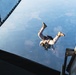A military parachutist has just exited the rear cargo ramp of a large military aircraft, visible in freefall against a high-altitude aerial view of agricultural terrain below. The jumper is wearing military camouflage uniform and combat boots, carrying equipment on their body. The photograph is taken from inside the aircraft looking down through the open ramp.