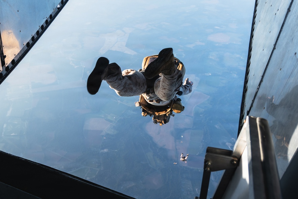 A military parachutist has just exited an aircraft and is in freefall, photographed from the aircraft's open ramp looking downward. The jumper is wearing military camouflage uniform and tactical gear with equipment pouches. The landscape below shows agricultural fields and terrain from high altitude.