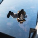 A military parachutist has just exited an aircraft and is in freefall, photographed from the aircraft's open ramp looking downward. The jumper is wearing military camouflage uniform and tactical gear with equipment pouches. The landscape below shows agricultural fields and terrain from high altitude.