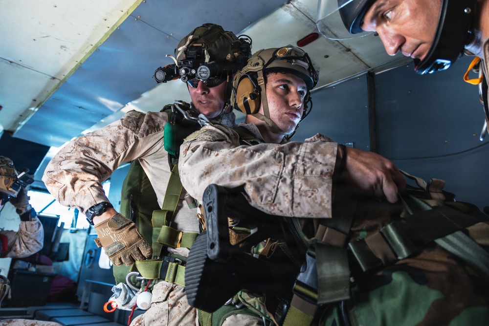 The image shows military personnel, likely U.S. Marines based on their MARPAT desert camouflage uniforms, inside what appears to be a military aircraft preparing for a parachute jump operation. At least two primary subjects are visible in full tactical gear including helmets, harnesses, and night vision devices. The scene suggests a HALO/HAHO or static-line jump preparation in progress.