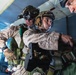 The image shows military personnel, likely U.S. Marines based on their MARPAT desert camouflage uniforms, inside what appears to be a military aircraft preparing for a parachute jump operation. At least two primary subjects are visible in full tactical gear including helmets, harnesses, and night vision devices. The scene suggests a HALO/HAHO or static-line jump preparation in progress.