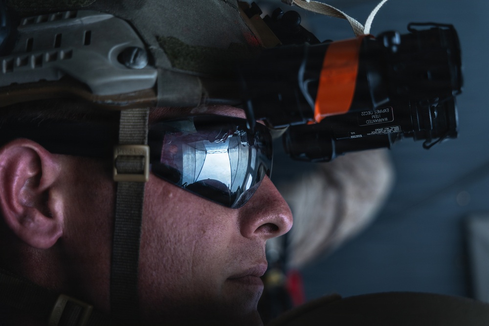 A close-up profile shot of a military personnel wearing tactical gear including a combat helmet and protective eyewear. A night vision or optical device is mounted on the helmet, secured with an orange strap, with a visible label reading 'WARRANTED ITEM EXPIRES DEC22'. The image is a high-contrast, dramatic photograph emphasizing the equipment and the subject's focused expression.