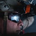 A close-up profile shot of a military personnel wearing tactical gear including a combat helmet and protective eyewear. A night vision or optical device is mounted on the helmet, secured with an orange strap, with a visible label reading 'WARRANTED ITEM EXPIRES DEC22'. The image is a high-contrast, dramatic photograph emphasizing the equipment and the subject's focused expression.