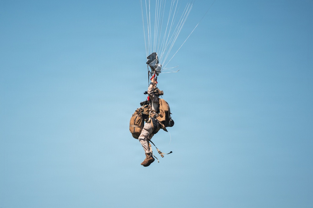 A military paratrooper is suspended mid-air beneath a partially deployed or collapsing parachute against a clear blue sky. The individual is heavily laden with tactical gear, including a large rucksack, equipment pouches, and what appears to be a weapon system. The parachute lines are visible extending upward to a canopy that appears to be in an irregular deployment state.
