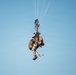 A military paratrooper is suspended mid-air beneath a partially deployed or collapsing parachute against a clear blue sky. The individual is heavily laden with tactical gear, including a large rucksack, equipment pouches, and what appears to be a weapon system. The parachute lines are visible extending upward to a canopy that appears to be in an irregular deployment state.
