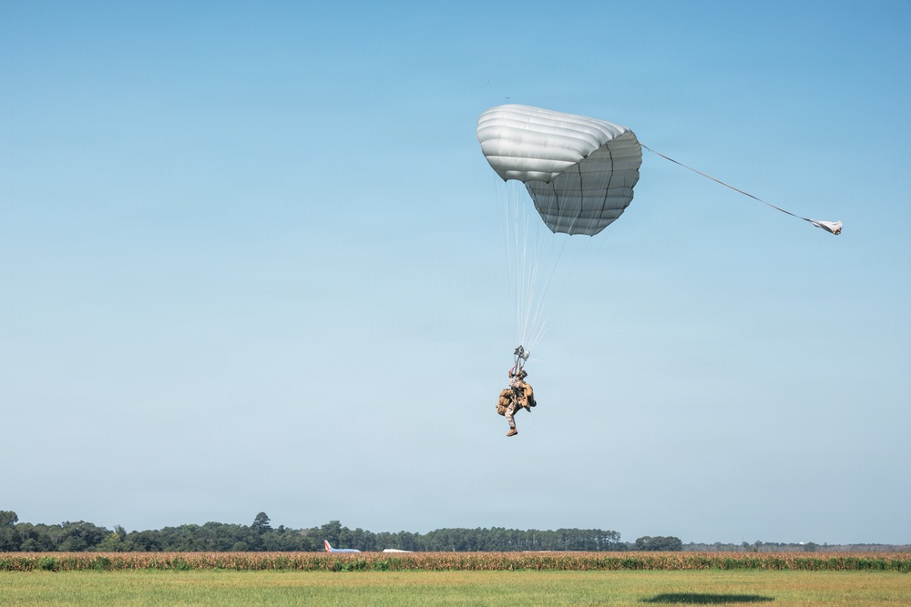 A military paratrooper is descending under a round, gray/silver parachute canopy over an open field near what appears to be an airfield. The jumper is equipped with full combat gear and a rucksack, and a static line or deployment cord is visible extending from the parachute. The background shows a tree line, agricultural fields, and what appears to be a small aircraft on the ground.