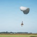 A military paratrooper is descending under a round, gray/silver parachute canopy over an open field near what appears to be an airfield. The jumper is equipped with full combat gear and a rucksack, and a static line or deployment cord is visible extending from the parachute. The background shows a tree line, agricultural fields, and what appears to be a small aircraft on the ground.