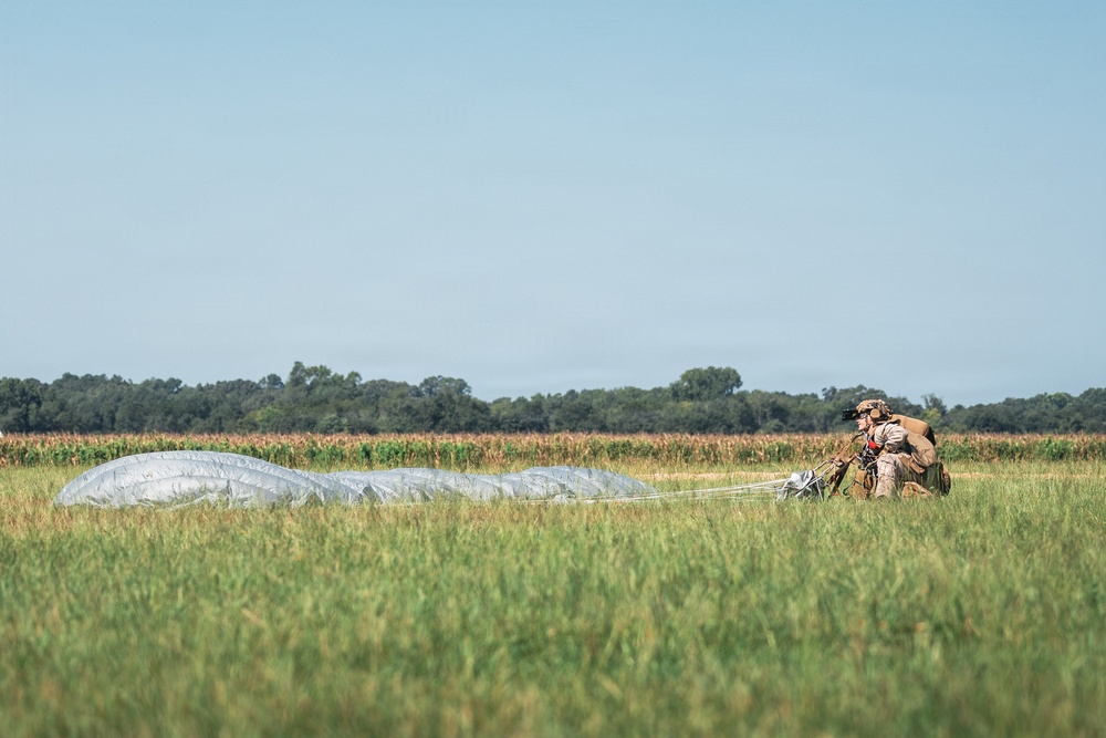 A military paratrooper has recently landed in an open grassy field and is in the process of gathering their parachute. The deflated silver/gray parachute canopy is spread across the grass to the left of the soldier. The setting appears to be a rural drop zone with a treeline and what appears to be a cornfield visible in the background.
