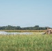 A military paratrooper has recently landed in an open grassy field and is in the process of gathering their parachute. The deflated silver/gray parachute canopy is spread across the grass to the left of the soldier. The setting appears to be a rural drop zone with a treeline and what appears to be a cornfield visible in the background.